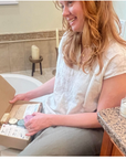 Woman sitting by a bathtub opening a self care subscription box with bath and wellness products.