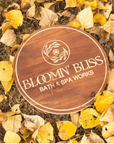 Wooden sign with 'Bloom'n Bliss Bath & Spa Works' logo on a bed of autumn leaves.