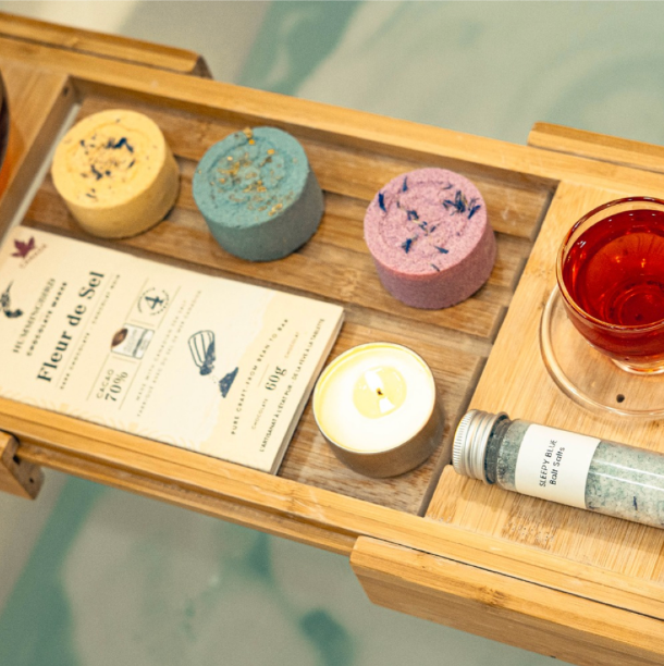 Contents of self care subscription box canada including massage candle, bath salts, chocolate, tea, and bath bombs over the bath water on bathtub tray.