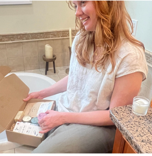 Woman sitting by a bathtub opening a self care subscription box with bath and wellness products.